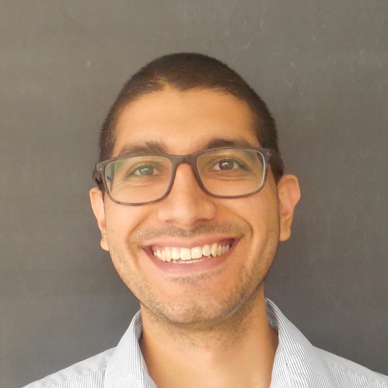 A man with short dark hair, glasses, and a trimmed beard smiles at the camera. He is wearing a light blue striped shirt and standing in front of a plain dark background.