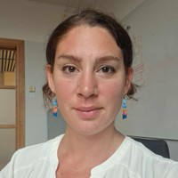 Woman with brown hair tied back, wearing colourful beaded earrings and a white blouse, sits in an office with a whiteboard and wooden door visible in the background.
