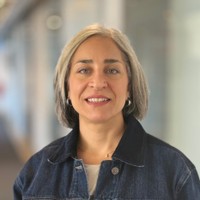 A woman with shoulder-length grey hair, wearing a dark denim jacket and silver hoop earrings, smiles softly whilst standing in a brightly lit corridor with glass walls.