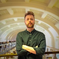 A man with fair hair and a beard stands indoors with arms crossed, in front of a large, arched ceiling with bright lighting and bookshelves in the background.