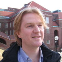 A man with light brown hair smiles outdoors in front of a large red-brick building with tiled roofs and arched walkways on a bright day.