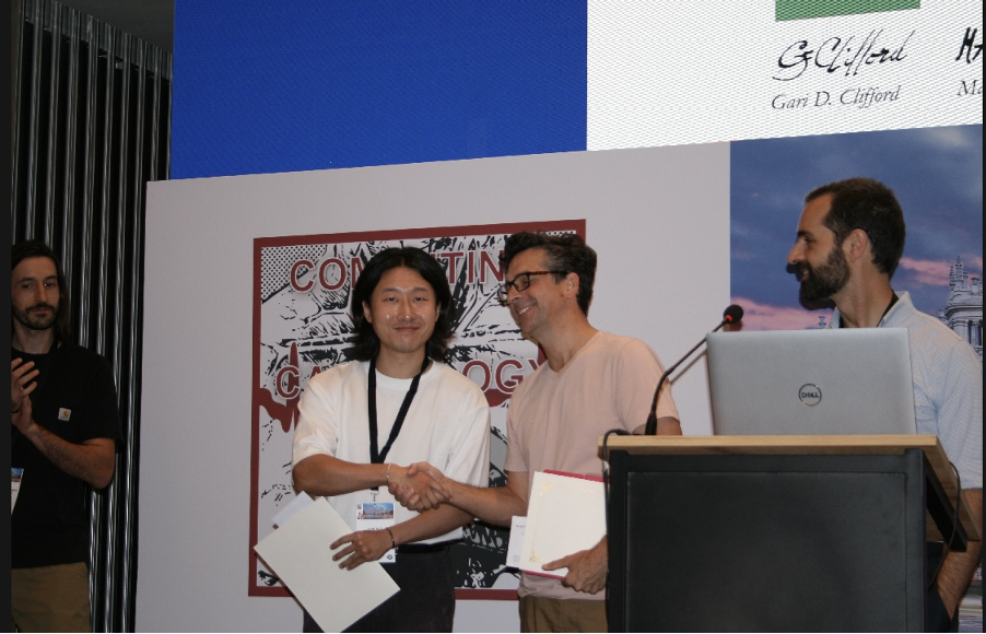 Two men shake hands on a stage while holding folders, with another man standing to the left and a laptop on a lectern to the right.