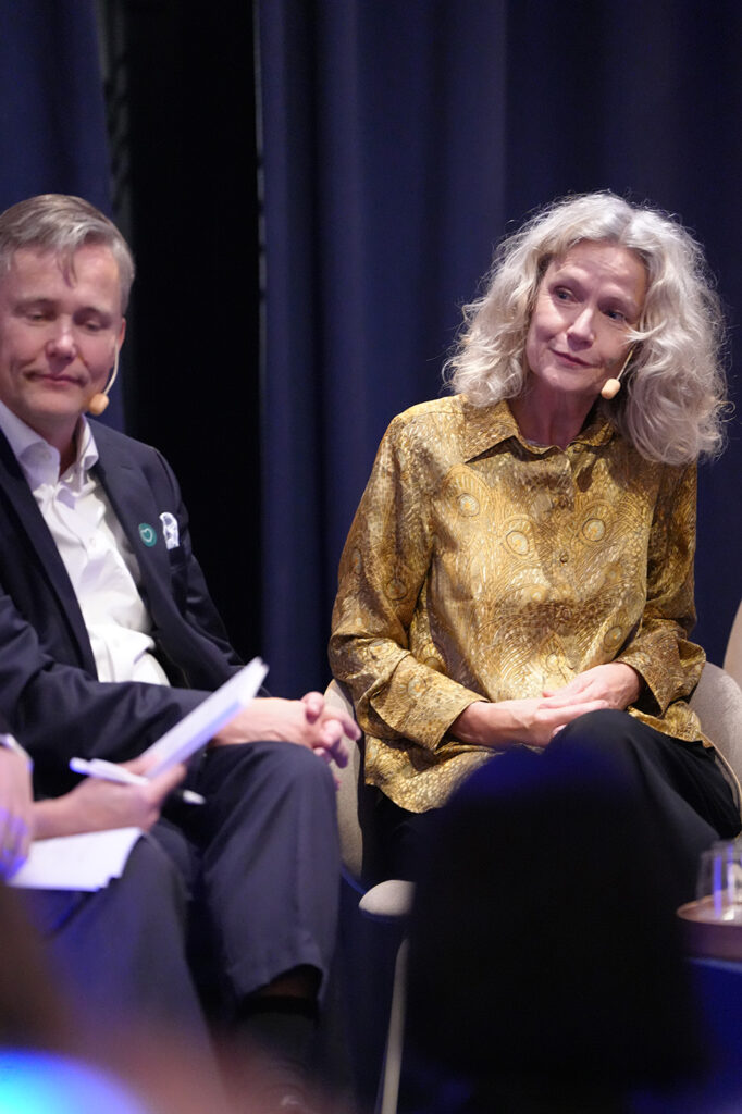 A man in a suit and a woman in a patterned gold blouse sit on stage during a panel discussion.