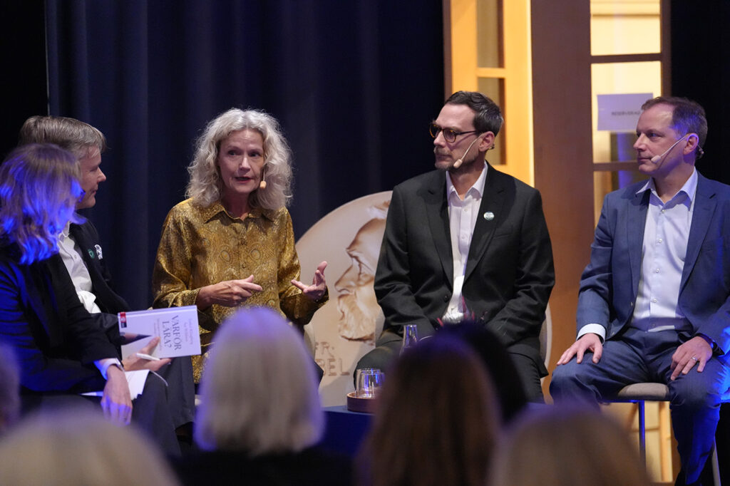 Four people seated on a stage during a panel discussion, with one woman holding a book titled Varför lära?.