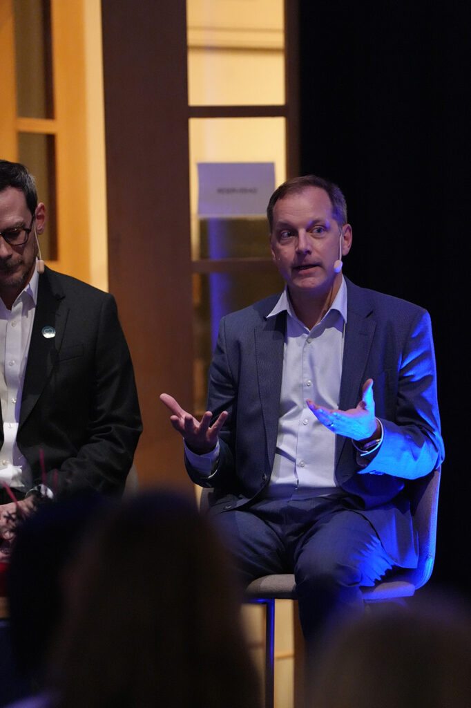 A man in a blue suit gesturing whilst speaking on a panel next to another man in a dark suit.