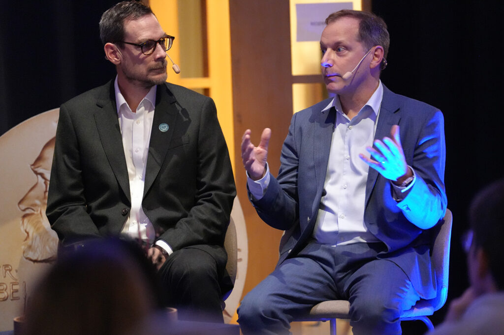 Two men in suits sitting on stage at an event, with one speaking and gesturing whilst the other listens.