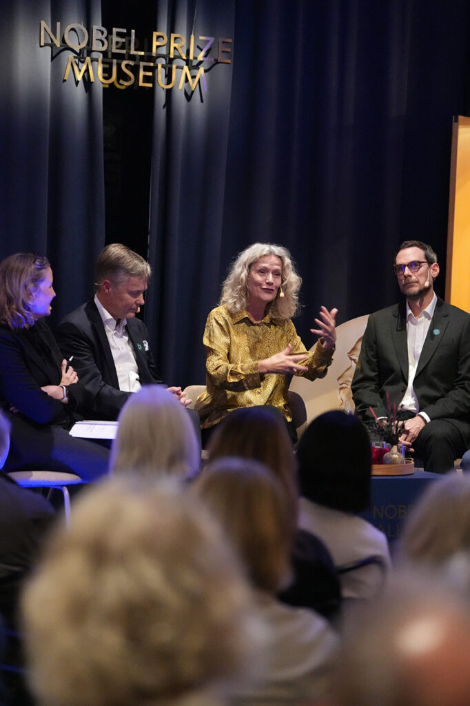 Four panellists sit on stage at the Nobel Prize Museum during a discussion, with a woman in a gold blouse speaking to the audience.