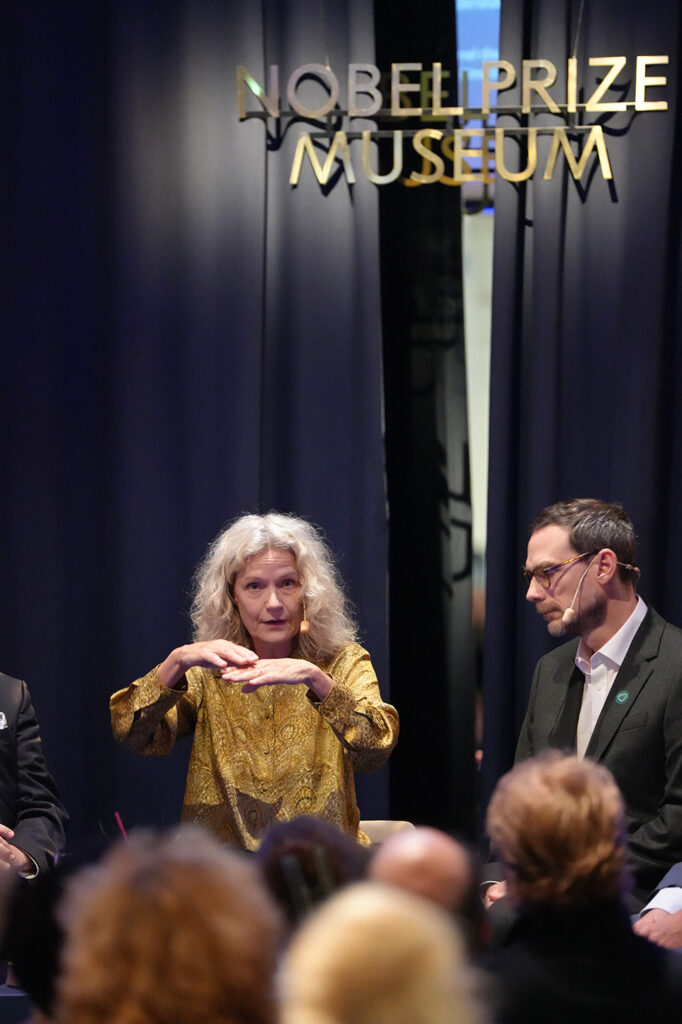 A woman in a gold patterned shirt gestures while speaking on a stage in front of a Nobel Prize Museum sign, seated next to a man in a suit.