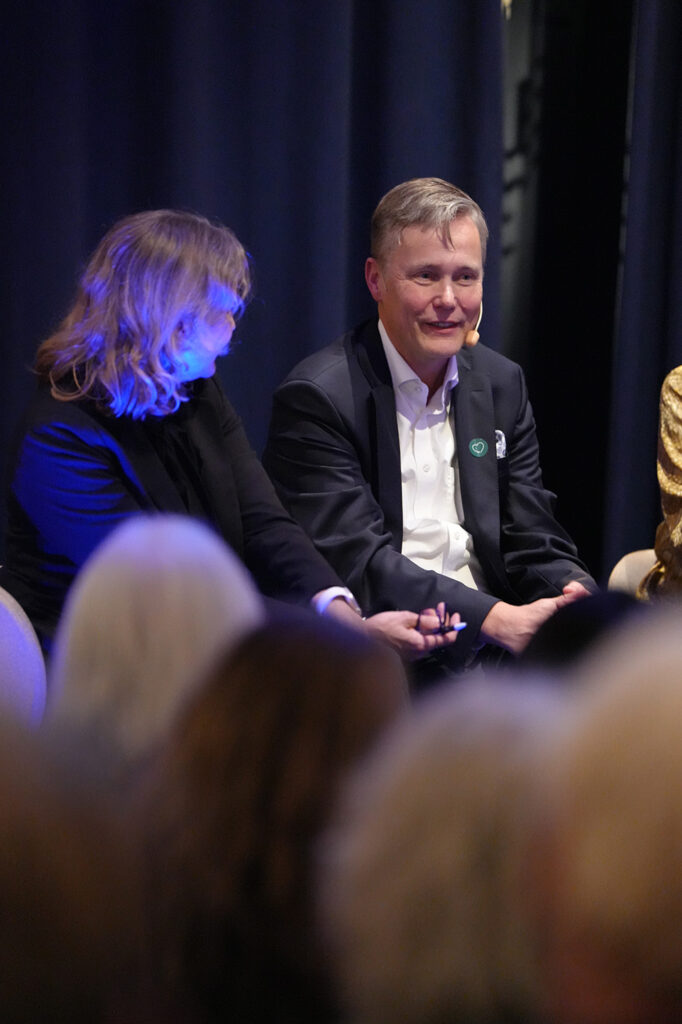 A man in a suit and a woman in a black jacket seated on a stage, speaking in front of an audience.