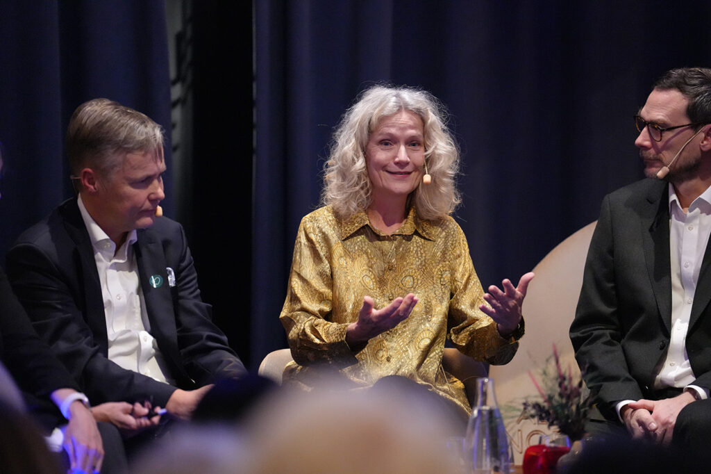 A panel of three people seated on stage, with a woman in a gold patterned blouse gesturing whilst speaking between two men in suits.