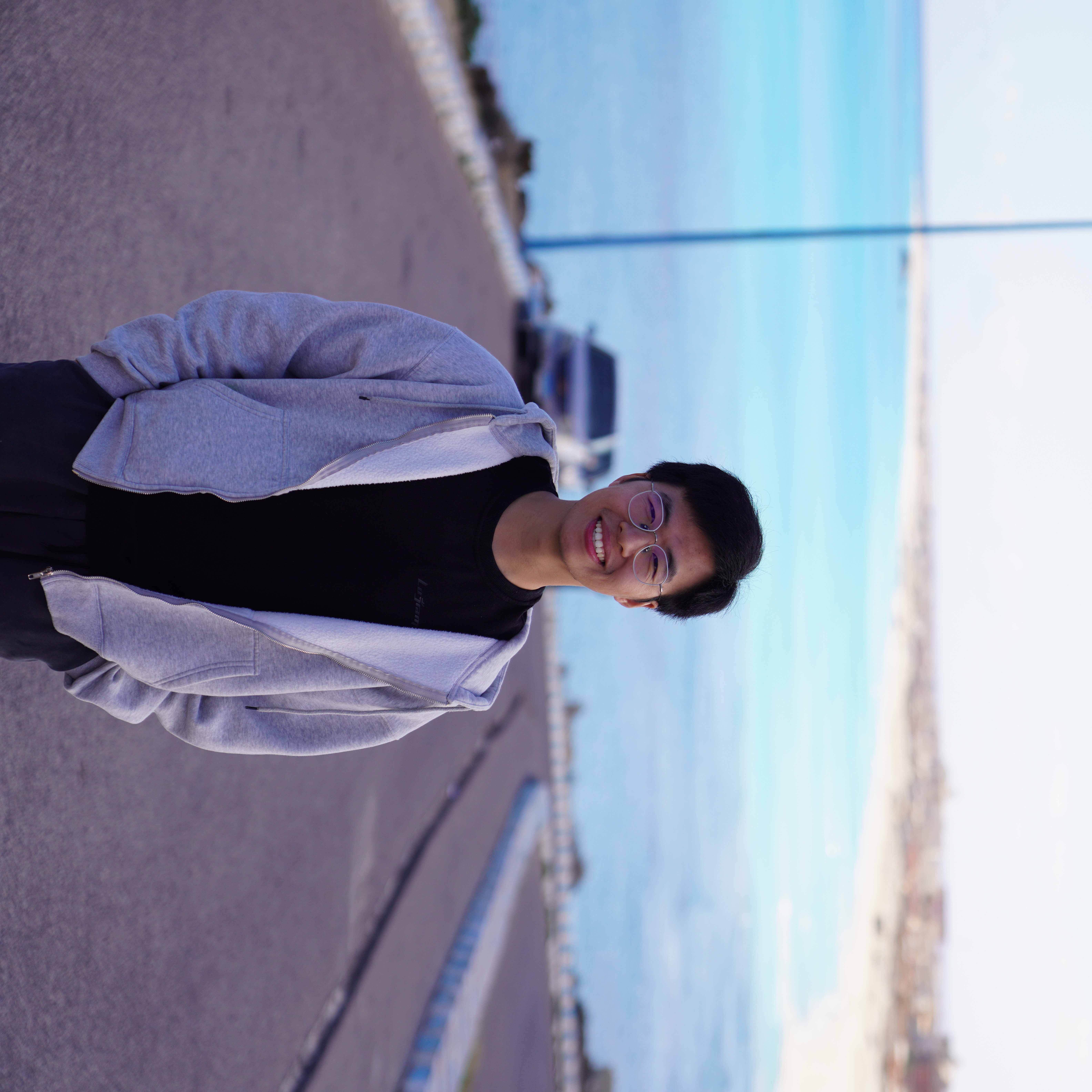 A young man in glasses and a grey hoodie stands on a paved road overlooking a blue sea and coastline.
