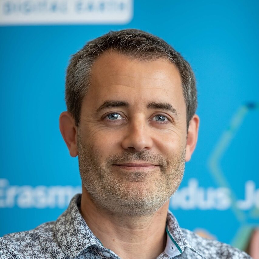 A man with short hair and light stubble smiling in front of a blue background.