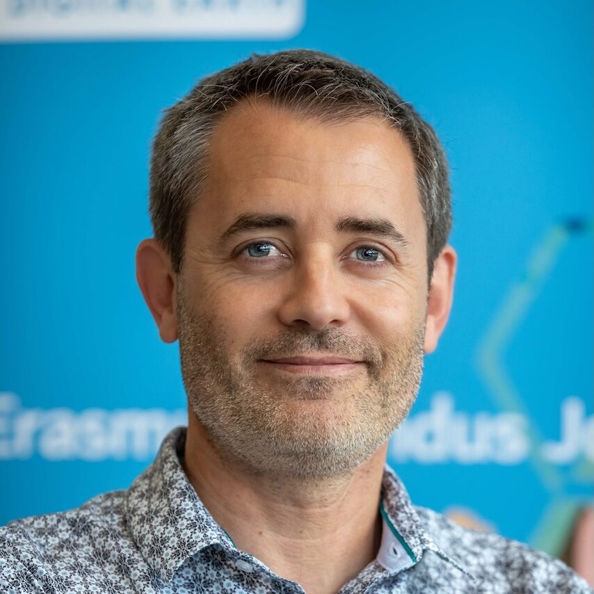 A man with short hair and light stubble smiling in front of a blue background.