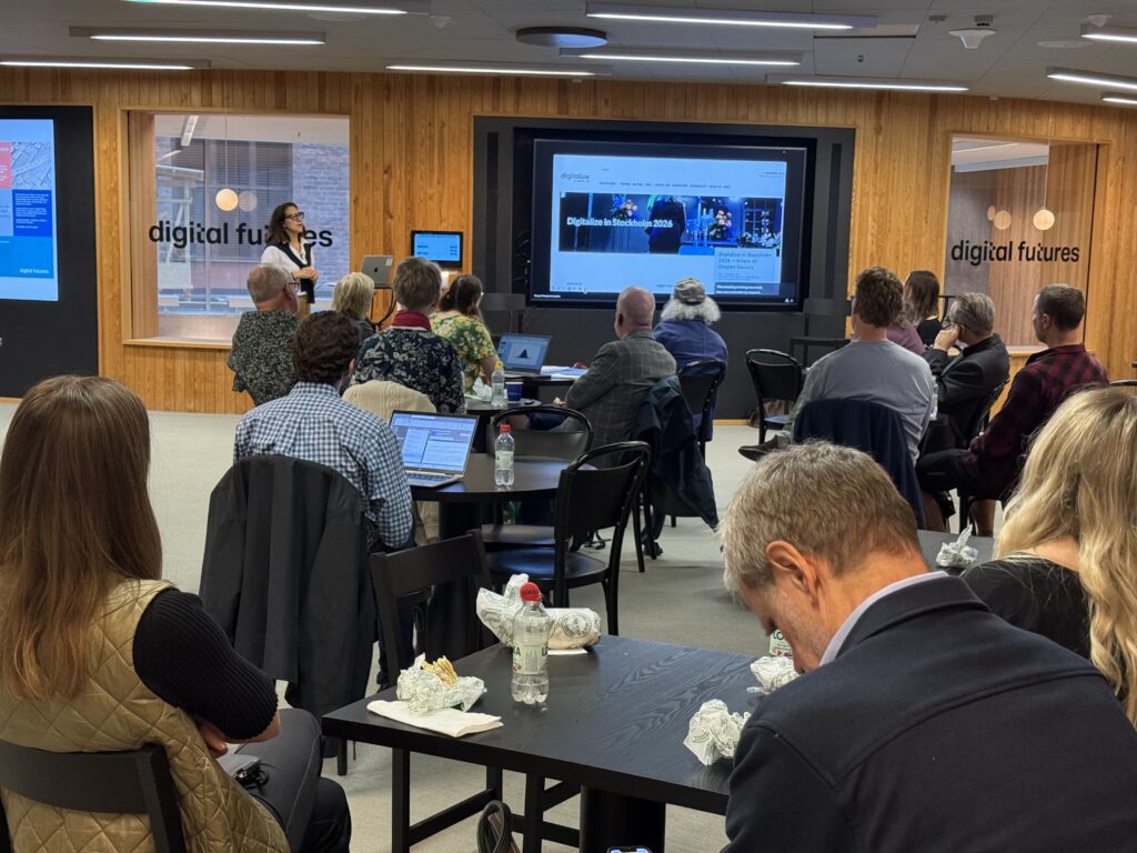 A woman stands at the front of a room presenting to an audience seated at tables facing a large screen displaying a Digital Futures website.
