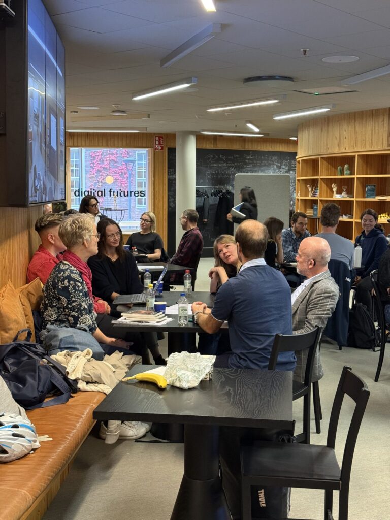 A group of people sit round a long table in a modern workspace, with some using laptops and others talking.