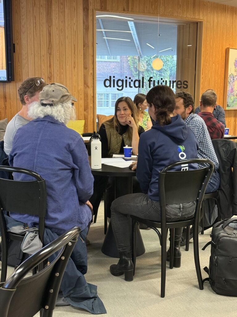 A group of people sit around a table for a meeting in an office with a digital futures sign on the glass wall.