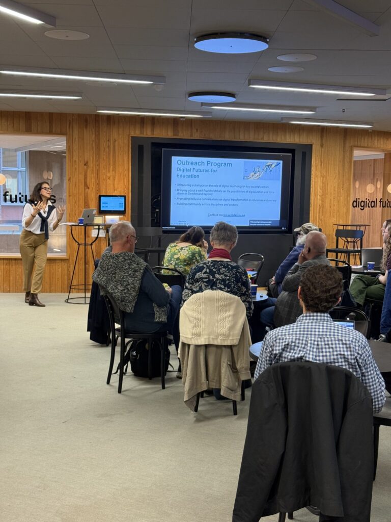 A presenter stands at the front of a room speaking to a seated audience facing a screen displaying a presentation titled Outreach Programme: Digital Futures for Education.