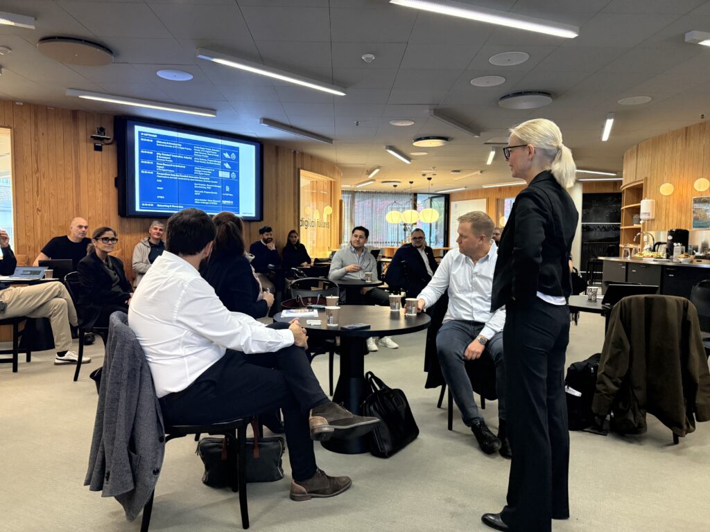 A professional woman stands and speaks to a group of people seated around tables during a meeting, with a presentation screen visible in the background.