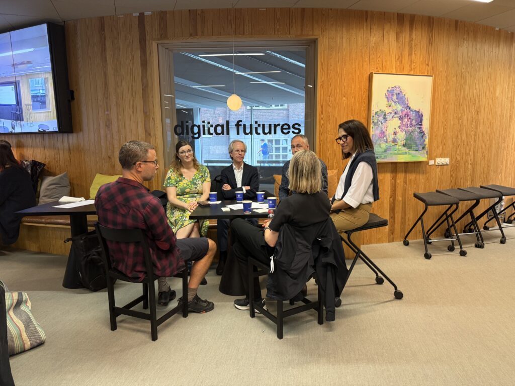 Five people sit round a table in a meeting room with wood-panelled walls and a window featuring the text digital futures.