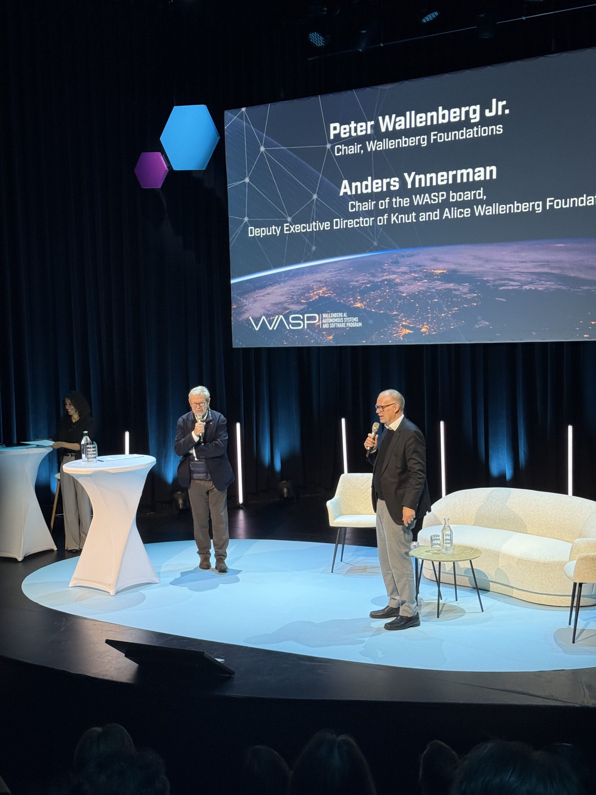 Two men standing on a stage during a presentation, with a large screen behind them displaying the names and titles of Peter Wallenberg Jr. and Anders Ynnerman.