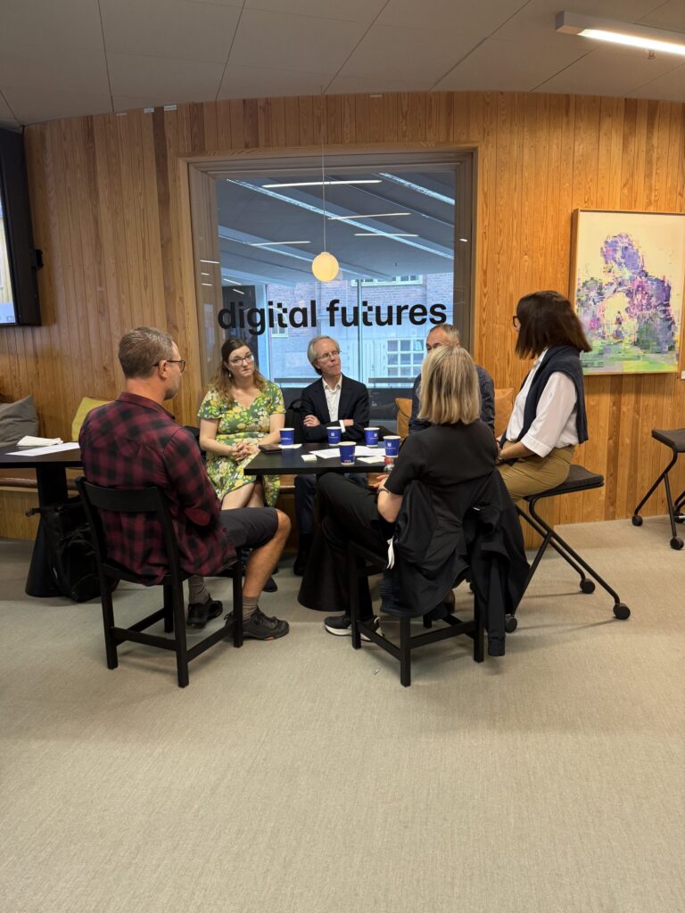 Four people sitting at a high table in an office, with digital futures printed on the window behind them.