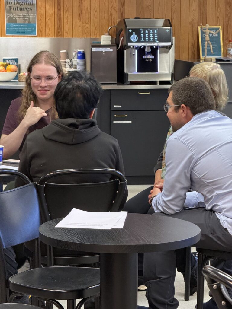Three people sit at a small round table in an office breakroom near a coffee machine.