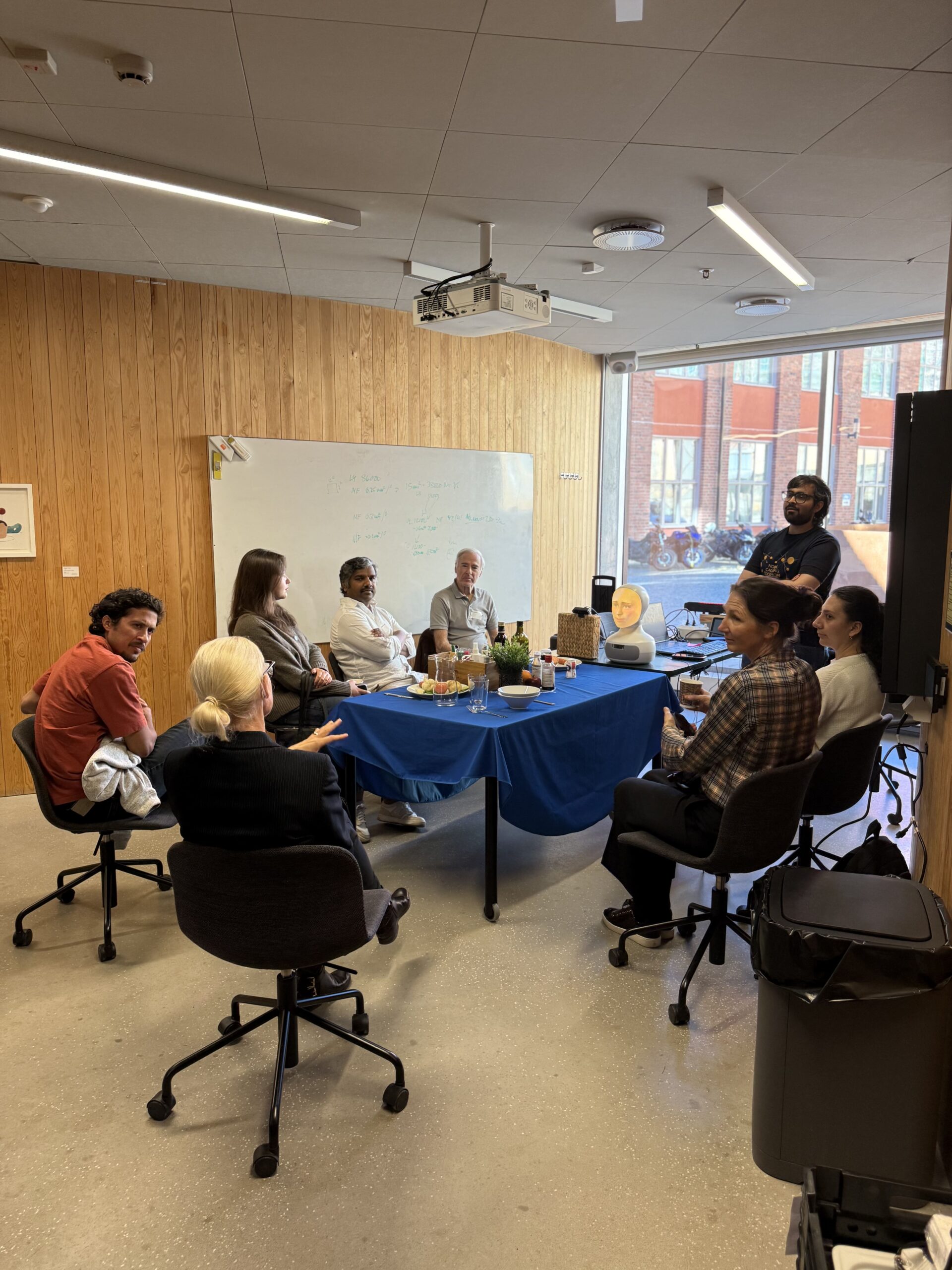 A group of seven people sitting around a table for a meeting in an office, with a small robot head placed on the table.