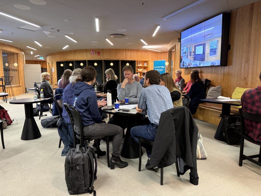 A group of people sit at round tables in a modern office space, talking and working during a meeting.