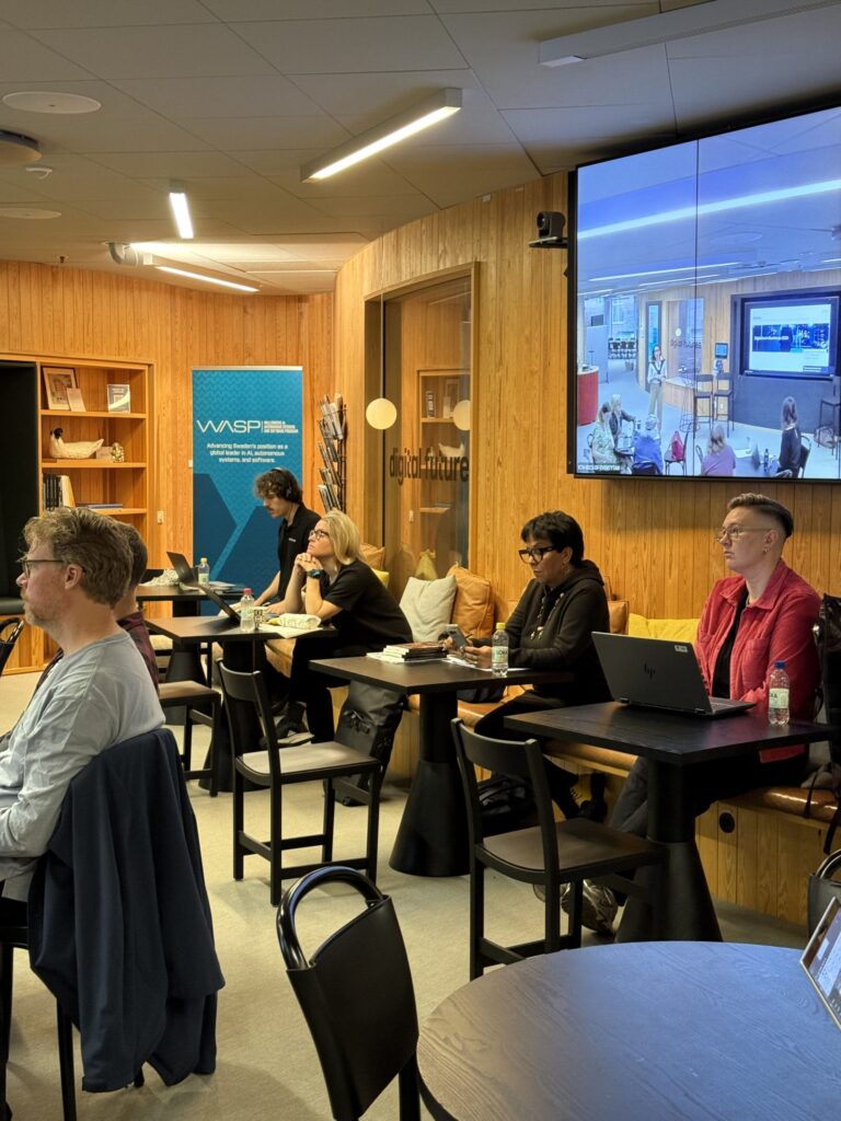 People working at laptops in a room with wood-panelled walls and a wall-mounted screen displaying a video call.
