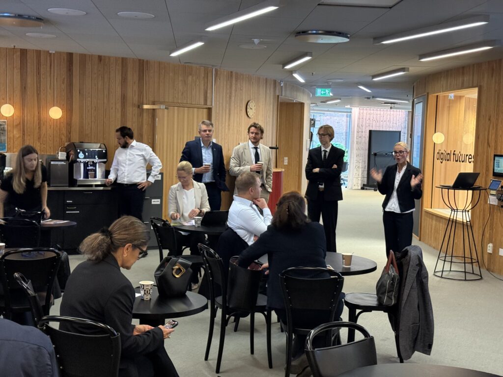 A group of people in business attire gather in a wood-panelled office space, with one woman standing and speaking to the room.