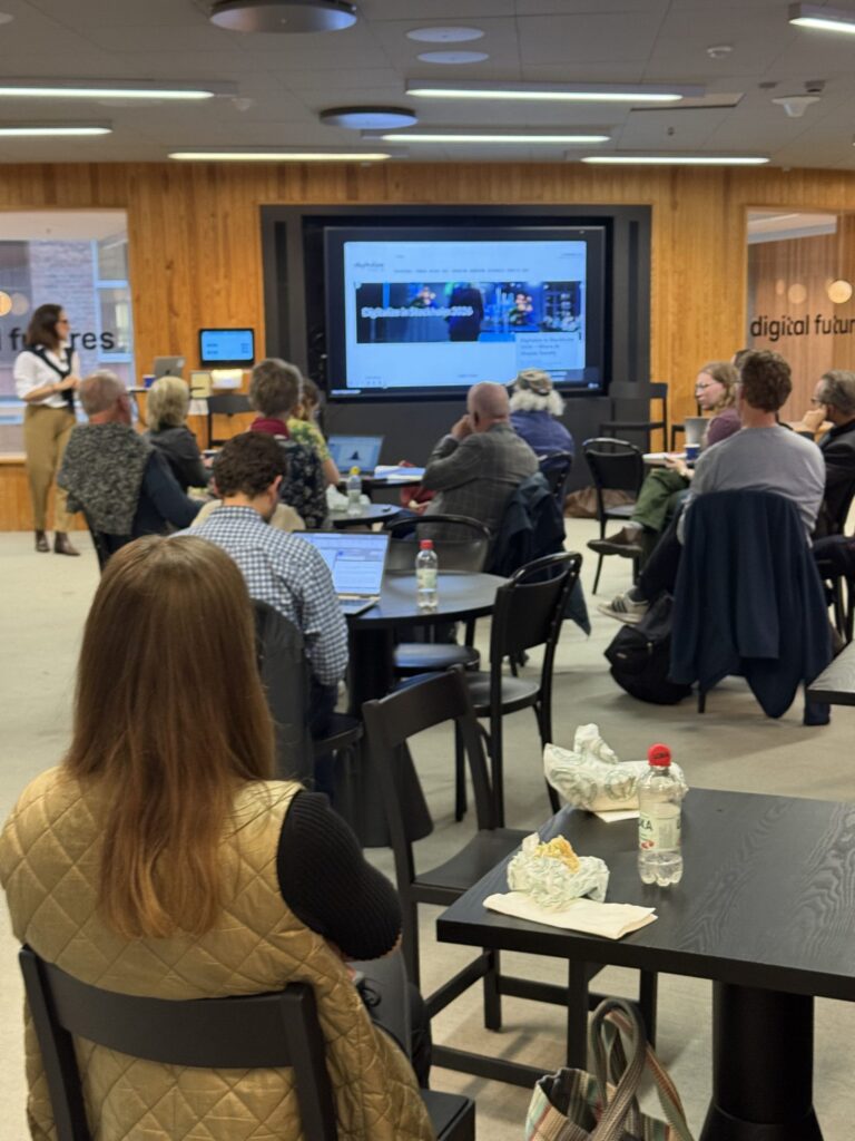 People seated at tables in a room, watching a presentation on a large screen.