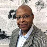 A man with glasses wearing a buttoned-down shirt and a patterned blazer poses in front of a wall with black and white line drawings.